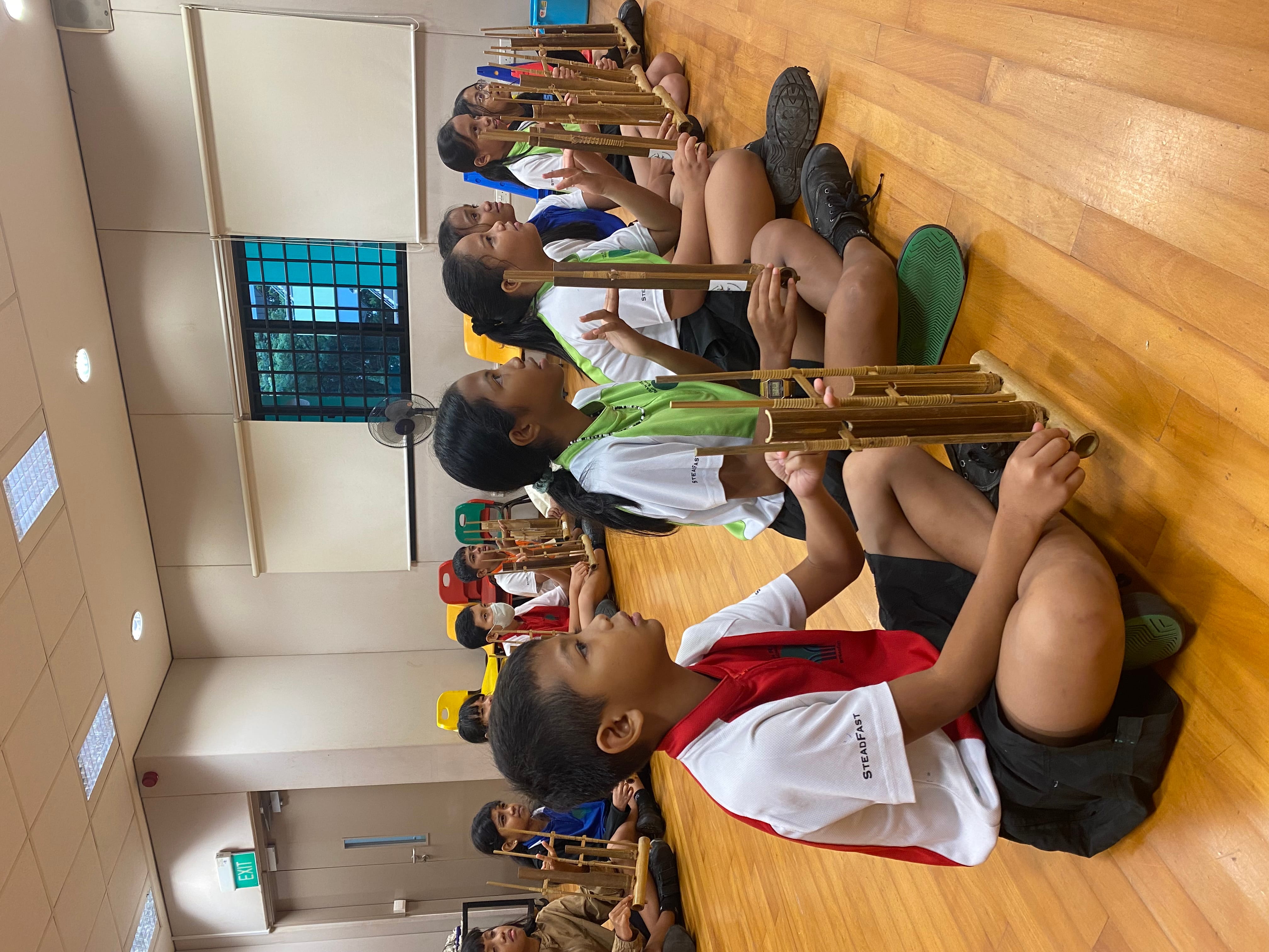 Children in school uniforms sit cross-legged on a wooden floor holding bamboo musical instruments during a class.